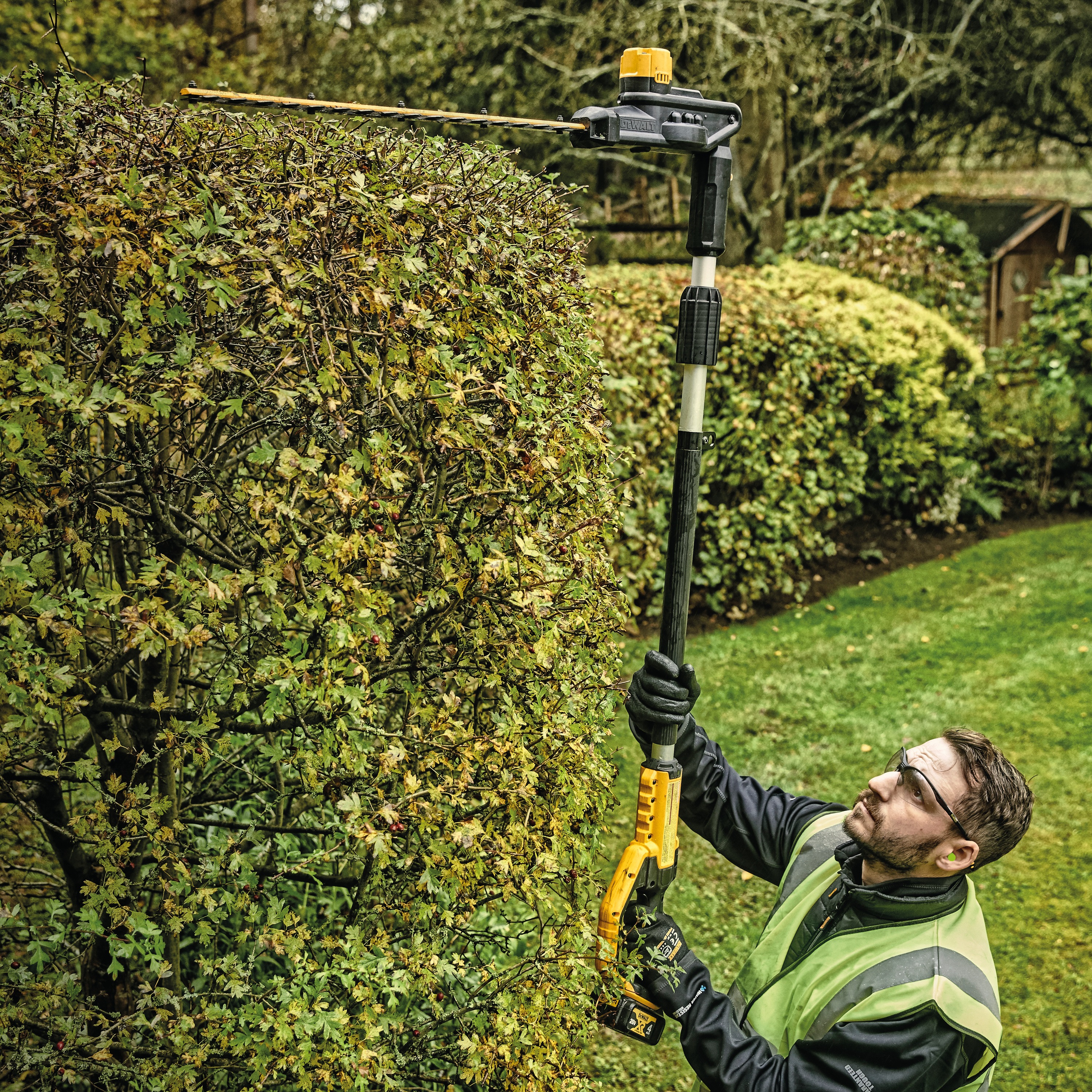 Overhead view of Pole Hedge Trimmer with battery being used by a person to cut through landscape outgrowth.