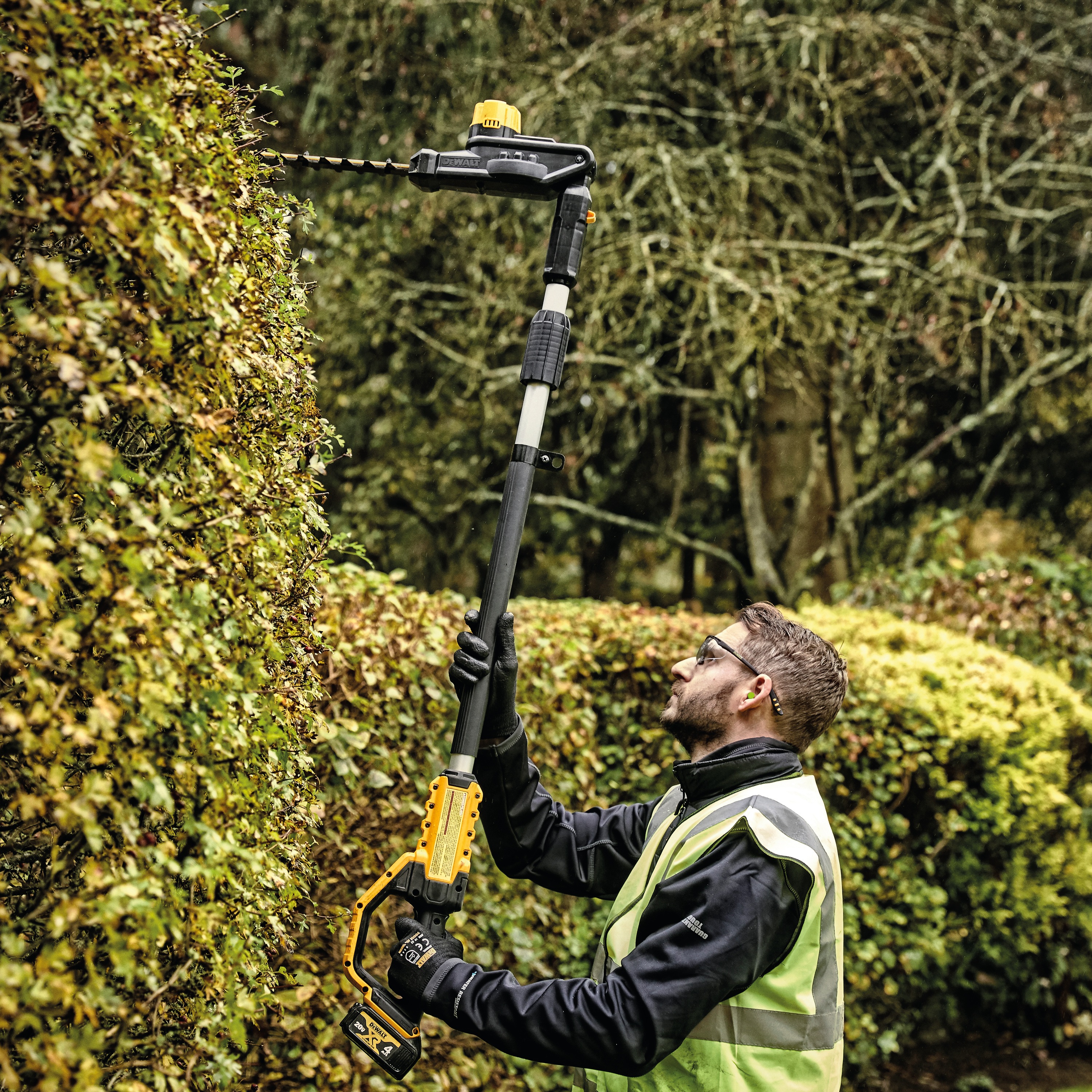 Pole Hedge Trimmer with a battery being used by a person to cut through landscape outgrowth.