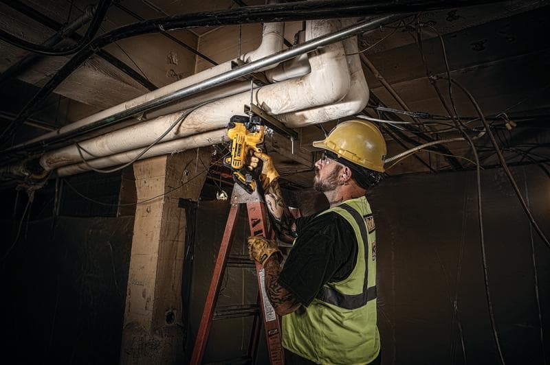 A construction worker wearing a safety vest and helmet is using a DEWALT pipe cutting tool (SKU: DCS350D1) to cut a metal pipe overhead while standing on a ladder in an industrial setting.