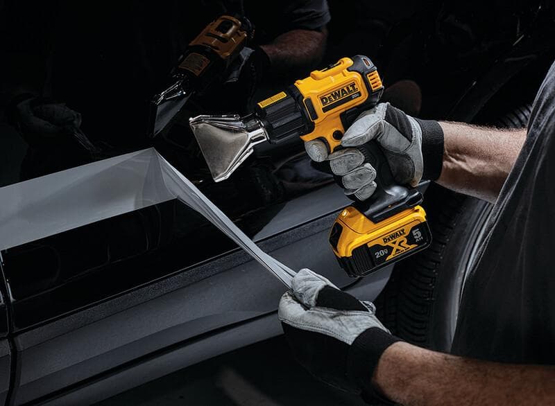 A person wearing safety gloves uses a DEWALT cordless heat gun to peel or apply a film or decal on a vehicle surface.