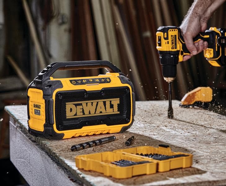 A DEWALT portable Bluetooth speaker is placed on a workbench next to a yellow tool case with drill bits. In the background, a person is using a DEWALT power drill to drill into the wooden surface, creating wood shavings. The scene takes place in a workshop environment.
