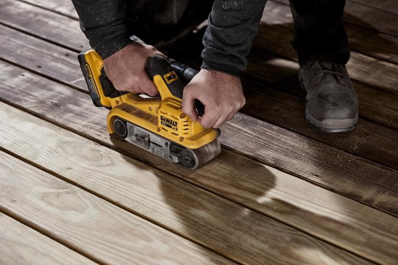 A person using a yellow DEWALT cordless belt sander to sand wooden deck boards.