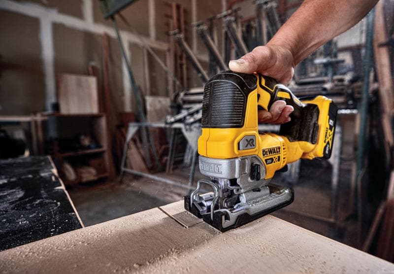 A close-up of a DEWALT cordless jigsaw being used to cut wood, with a person's hand holding the tool in a workshop setting.