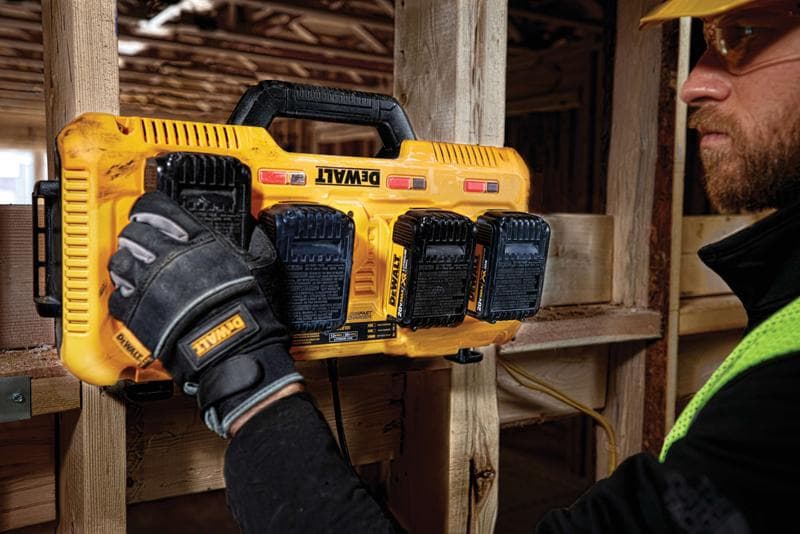 A Dewalt DCB104 yellow multiport battery charger mounted on a wooden beam at a construction site. A person wearing Dewalt-branded gloves is handling the charger, and there are four Dewalt batteries attached to it.