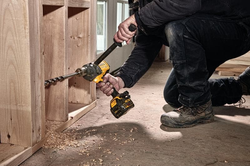 A person using a DEWALT cordless drill with an auger bit to drill a hole in a wooden structure, with wood shavings scattered on the floor.
