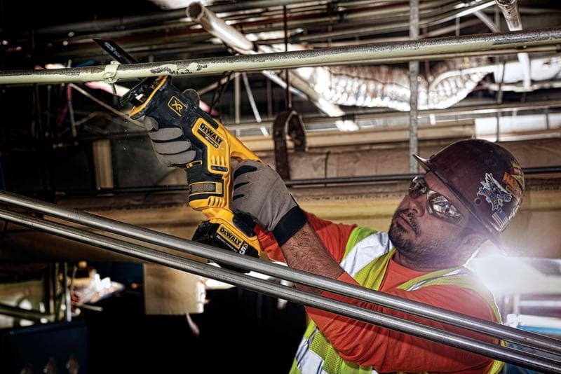 A person wearing work gloves, an orange shirt, safety vest, and helmet is using a DEWALT DCS367B cordless reciprocating saw to cut a metal pipe in an industrial setting. The person's face is blurred for privacy.