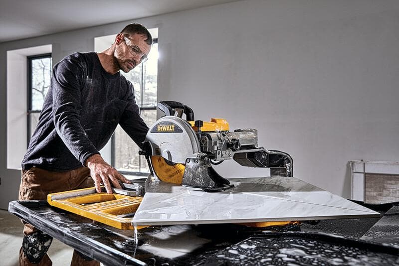 A person operating a DEWALT tile saw (SKU: D36000S) to cut a large piece of tile in a well-lit indoor workspace.