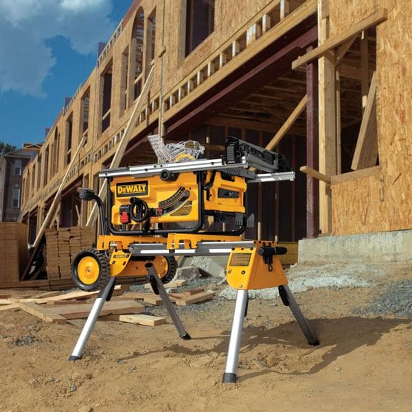 A DEWALT DW7440RS portable table saw stand with a yellow and black table saw set up outdoors at a construction site, where a wooden framed building is being constructed.