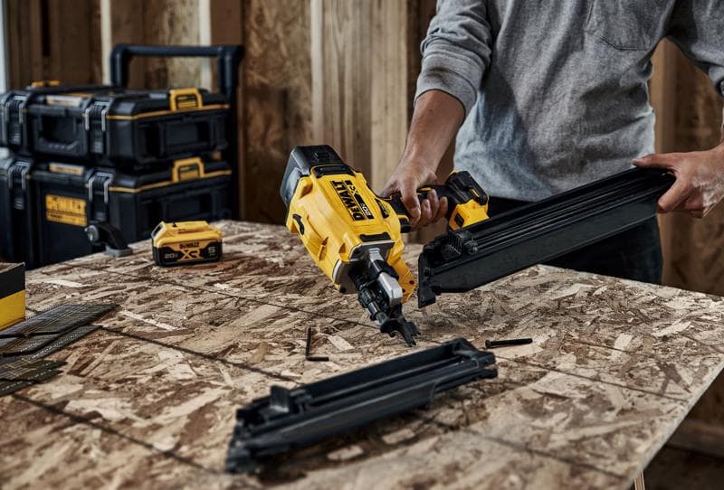 A person assembling a yellow DEWALT nail gun at a workbench in a woodworking workshop. The table is scattered with nails and nail gun components, and DEWALT toolboxes are visible in the background.