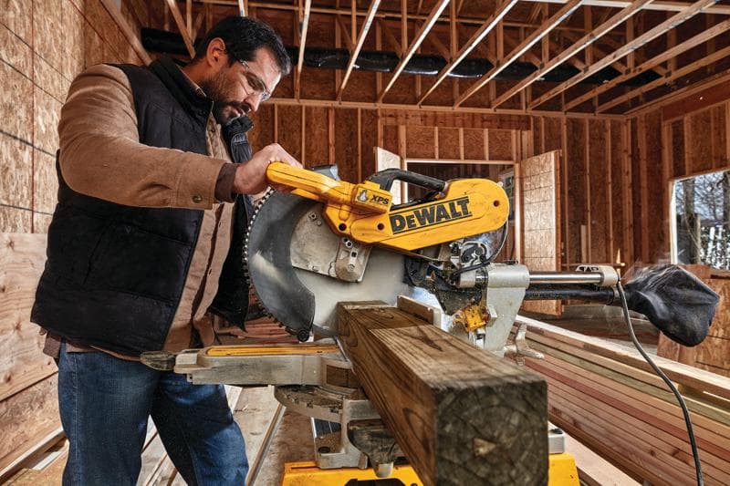 A person operating a DEWALT DWS780 miter saw in a construction site, cutting a wooden beam inside a partially built structure.