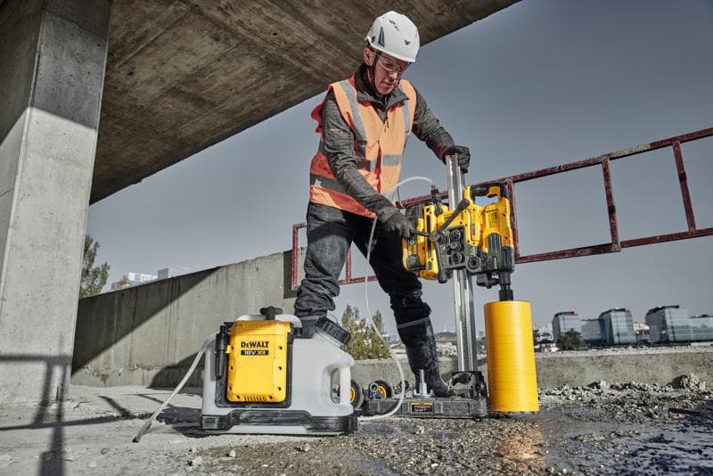 A worker wearing a helmet and safety vest operates a DEWALT DCPS151 power tool with a cylindrical yellow attachment, drilling into concrete at a construction site. A DEWALT water supply unit is placed next to the tool.