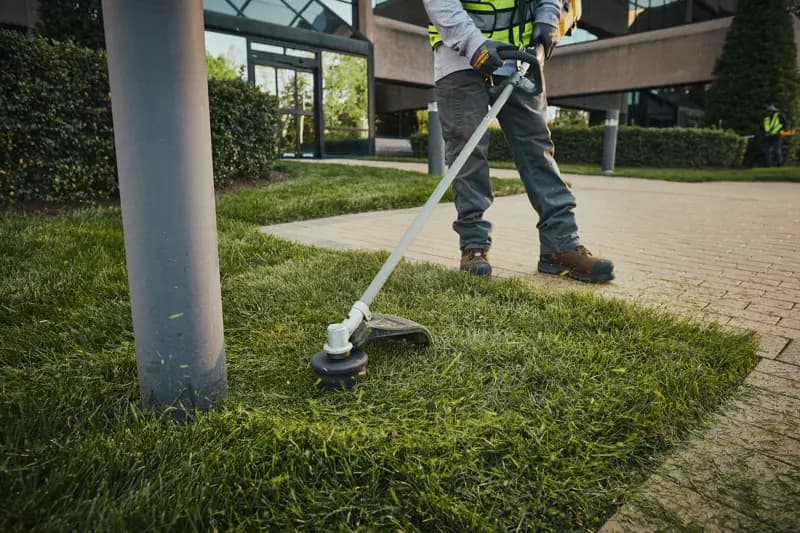 A person using a DEWALT string trimmer to edge grass along a sidewalk in a landscaped outdoor area, wearing gloves and work boots.