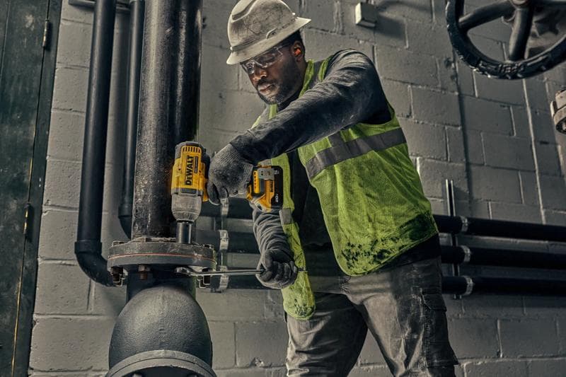 A worker wearing a hard hat and high-visibility vest is using a DEWALT cordless impact wrench (model DCF900P1) to tighten bolts on a large industrial pipe in an indoor setting.