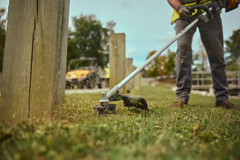 A person using a DEWALT cordless string trimmer to cut grass near wooden posts outdoors. The tool is actively cutting grass with clippings flying.