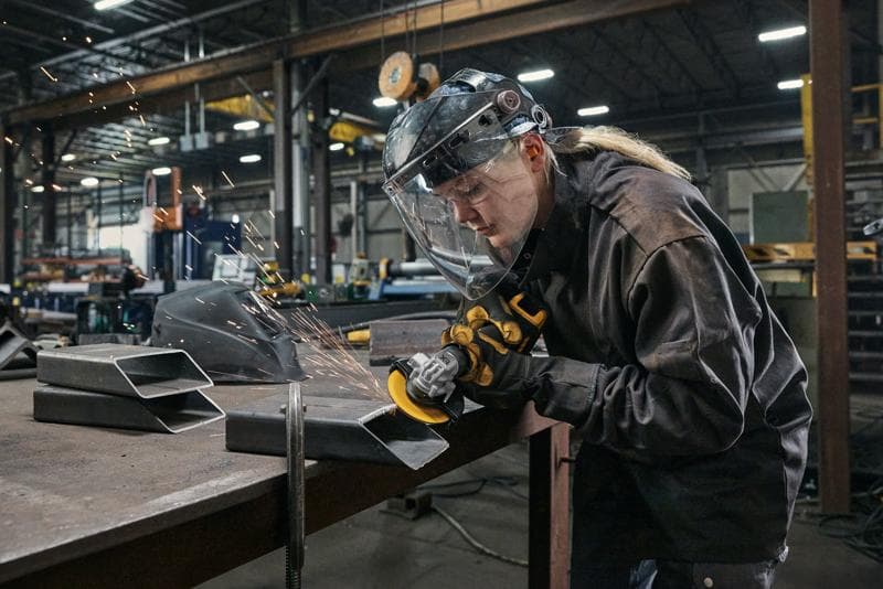 A worker wearing protective gear, including a face shield and gloves, uses a DEWALT angle grinder to cut or grind metal pieces in an industrial workshop. Sparks are flying from the grinder.