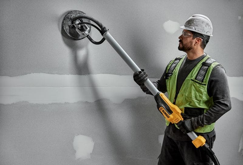 A construction worker wearing a hard hat and safety vest uses a DEWALT DWE7800 drywall sander with a telescopic handle to sand a gray wall.
