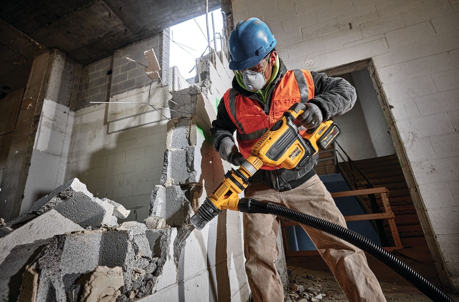 A construction worker wearing a blue hard hat and orange safety vest uses a DEWALT DCH614B cordless rotary hammer to demolish a concrete wall, with debris falling around and a vacuum hose attached to the tool.