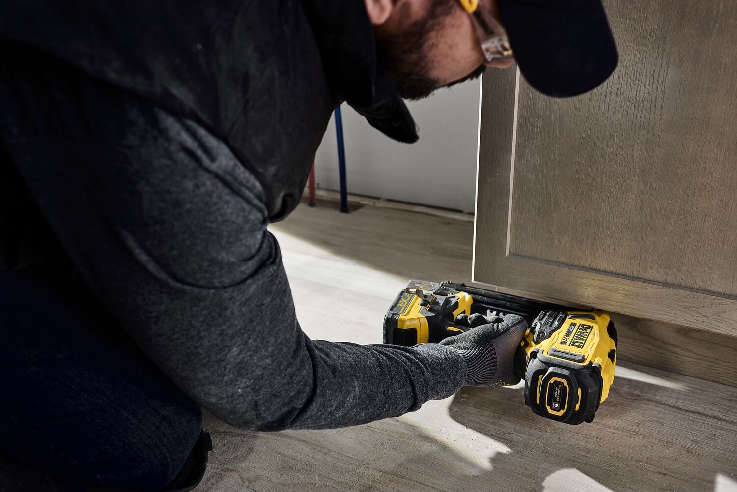 A person using a DEWALT DCN210 power tool to work on the bottom of a wooden cabinet, likely performing construction or installation tasks on a floor.