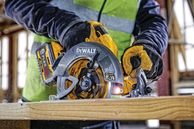 A close-up view of a person using a DEWALT 60V FLEXVOLT cordless circular saw to cut through a piece of wood. The person is wearing work gloves and a high-visibility vest inside a construction site.
