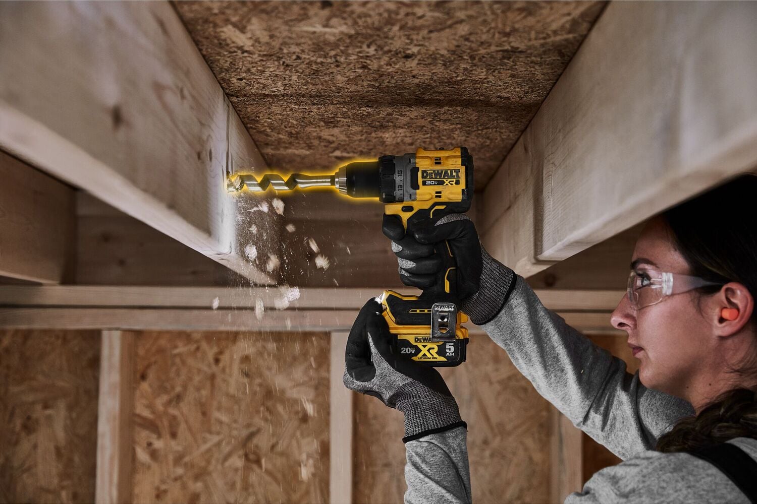 A person using a DEWALT cordless drill (model DCD800B) to drill a hole in a wooden beam, with wood shavings falling. The person is wearing gloves and their face is blurred.