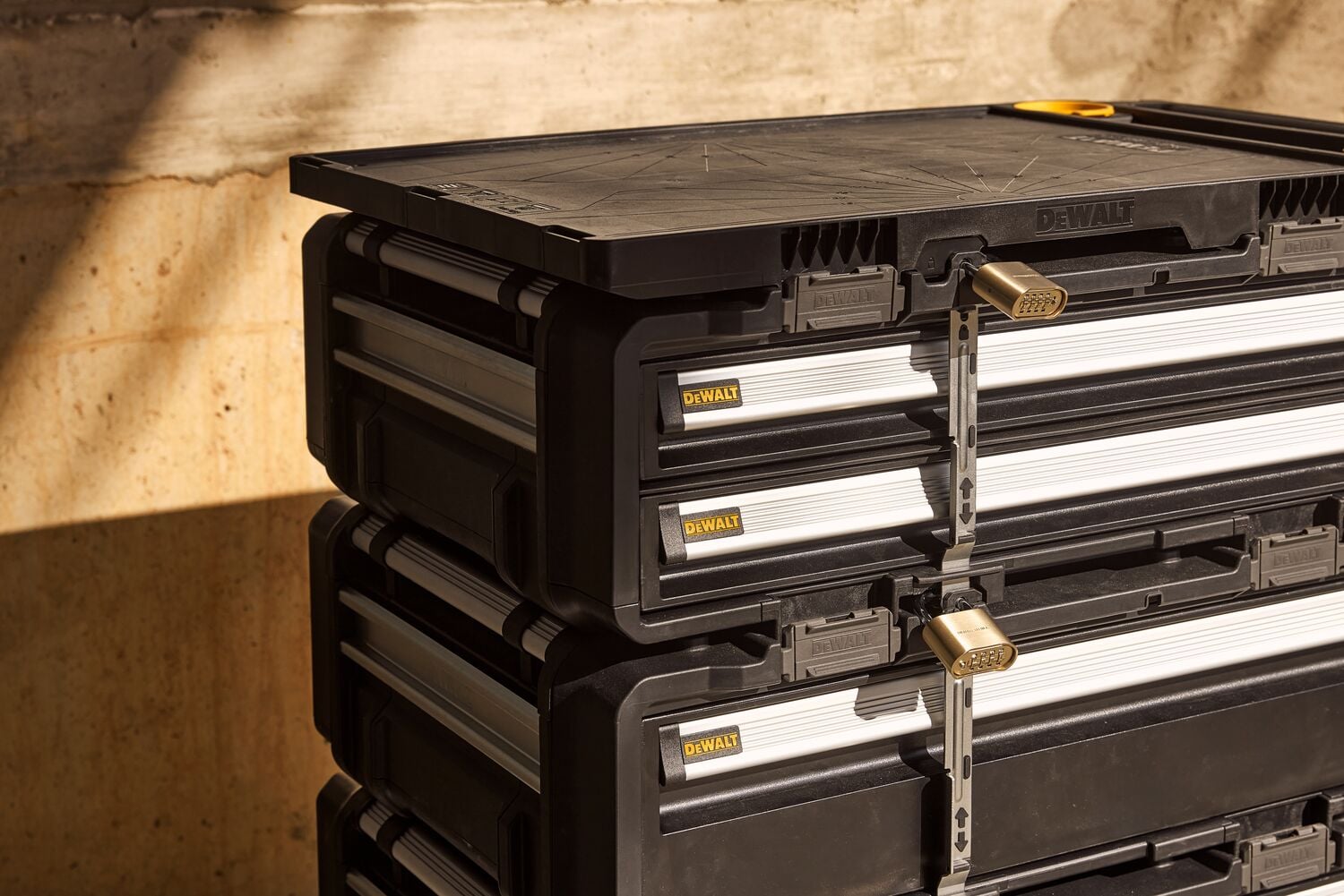 Stacked DEWALT lockable tool storage boxes, made of black plastic with metal accents, placed against a wall in sunlight.