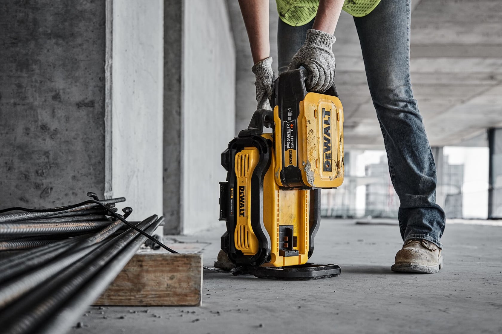A person wearing gloves and work boots is handling a portable yellow DEWALT power tool at a construction site. Rebar and concrete are visible nearby.