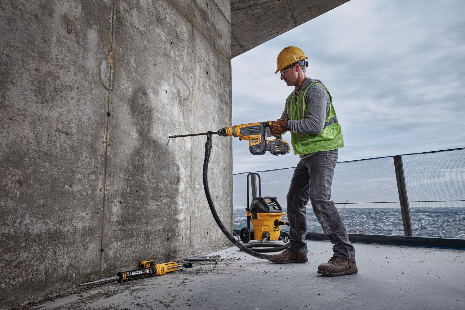 A construction worker wearing a yellow hard hat and green safety vest uses a DEWALT cordless rotary hammer drill to drill into a concrete wall. The tool is connected to a dust extractor through a hose, and other DEWALT equipment is visible on the ground nearby.