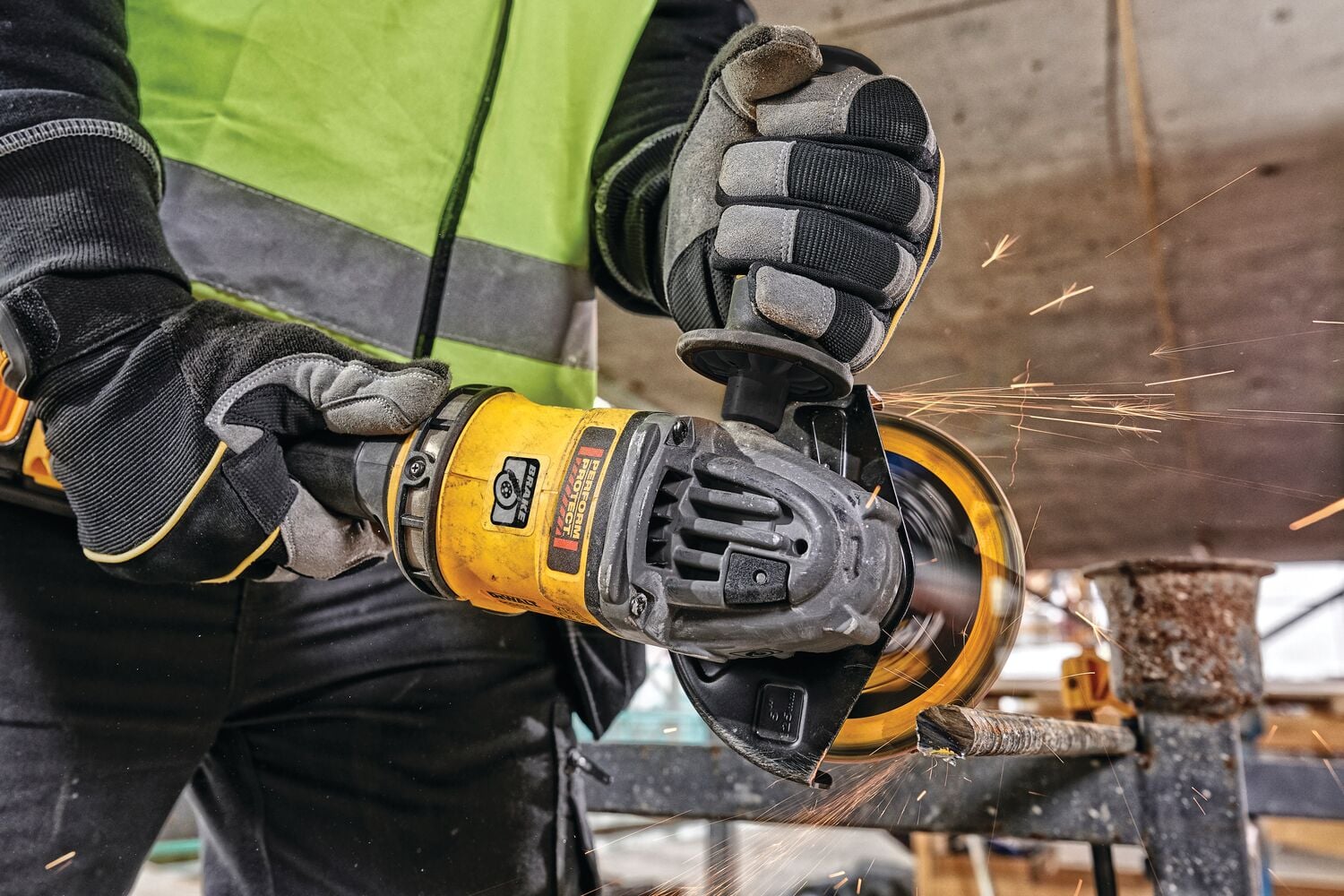 Close-up of a person wearing gloves and a high-visibility jacket, using a yellow DEWALT cordless angle grinder to cut metal, with sparks flying.
