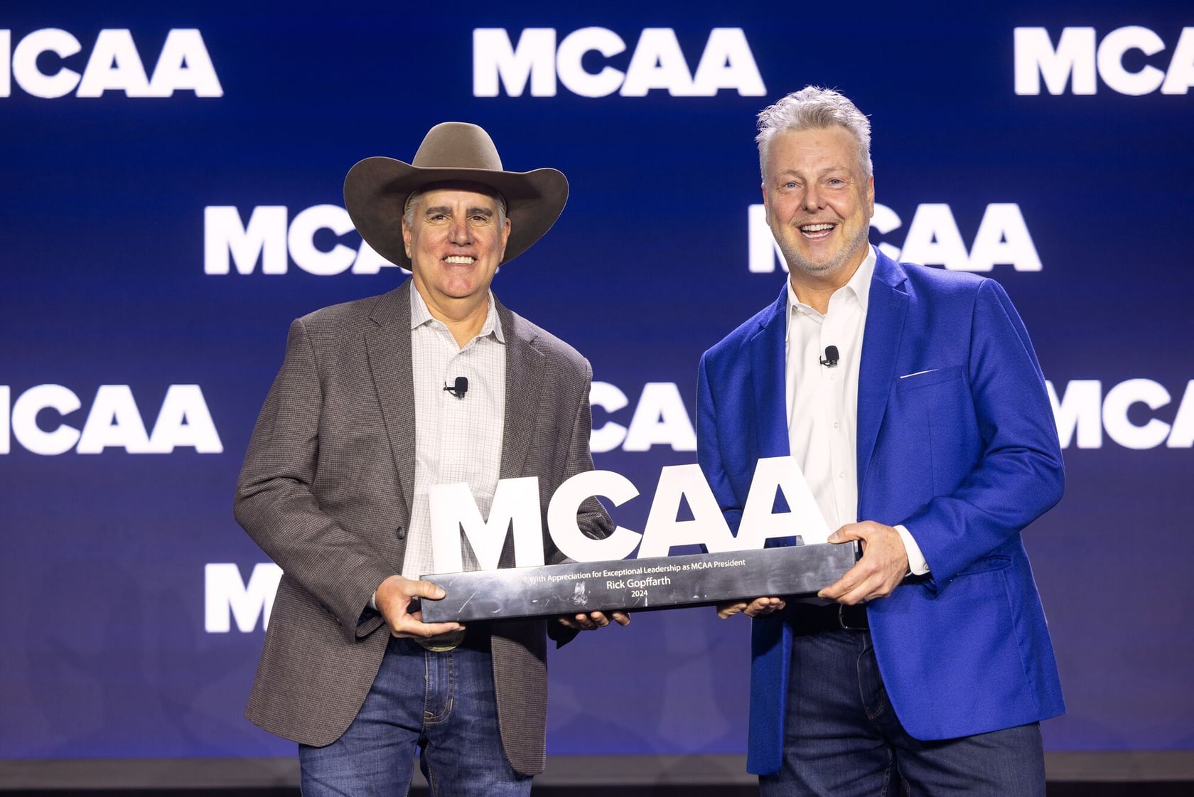 A person wearing a light-colored blazer is speaking into a microphone onstage in front of a screen showing images and text, including group photos with people wearing yellow shirts.