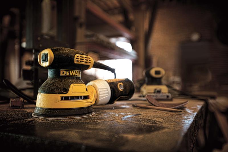 A DEWALT DWE6421 yellow and black electric orbital sander is placed on a dusty workbench in a workshop. There are additional sanding tools and sandpaper nearby in the background.