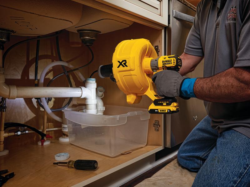 A person is using a DEWALT XR cordless drain cleaner under a kitchen sink, working on plumbing pipes. The tool is yellow and black, and a plastic container is placed beneath the pipes to catch any water. Additional tools are seen nearby.
