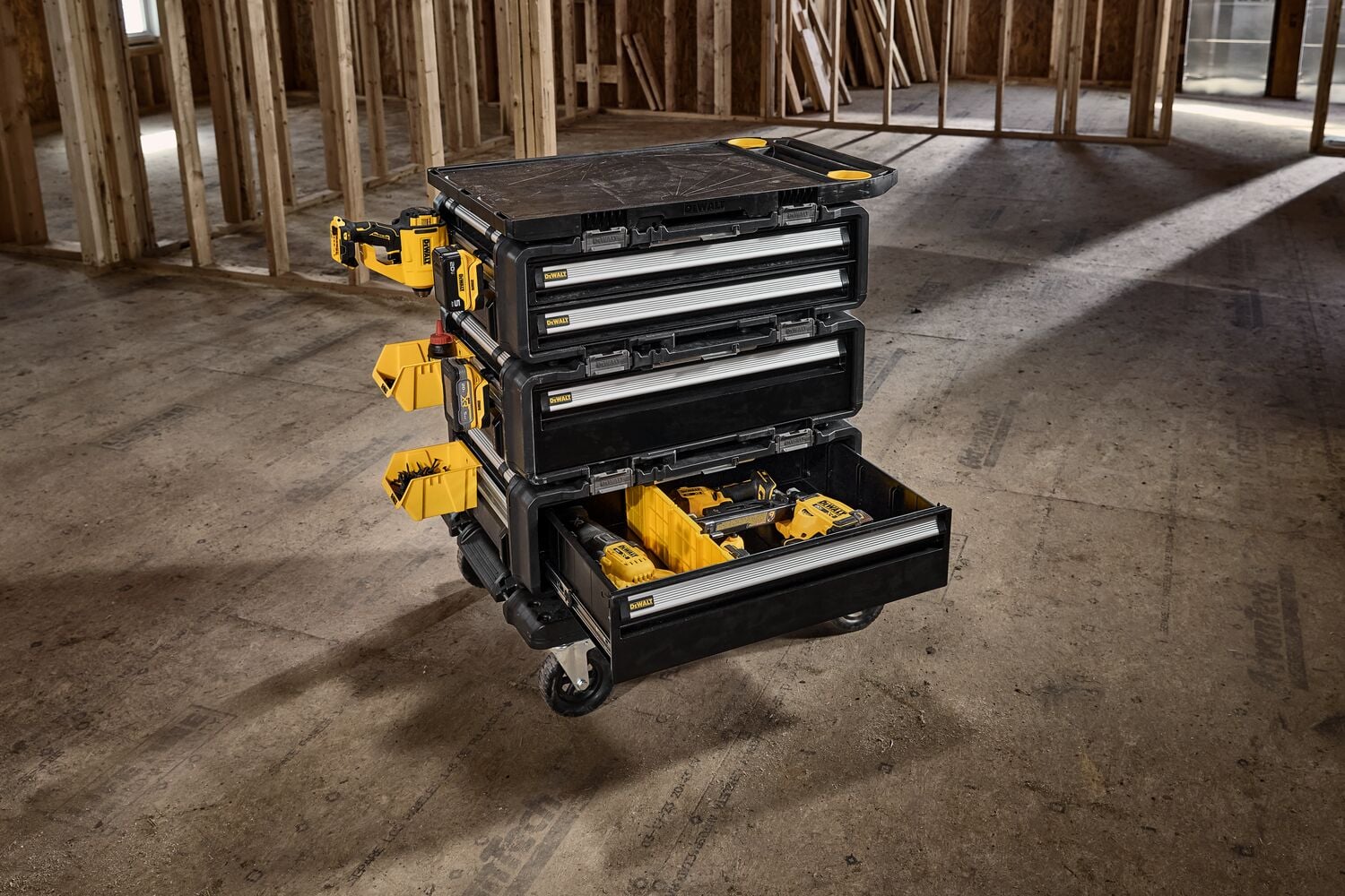 A DEWALT mobile tool storage cart with multiple drawers open, containing yellow compartments and tools, positioned in an unfinished construction site with wooden framing.