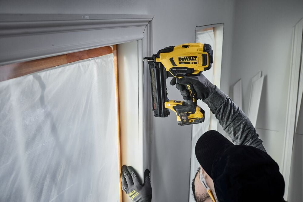 A person wearing gloves and safety glasses uses a yellow DEWALT nail gun (model DCN210) to fasten trim around a window. The trim and window are partially covered for protection during installation.