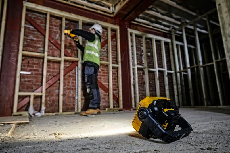 A DEWALT portable work light (SKU: DCL077) sits on the floor of a construction site, illuminating the area. In the background, a construction worker wearing safety gear is working near a wooden frame against a brick wall.
