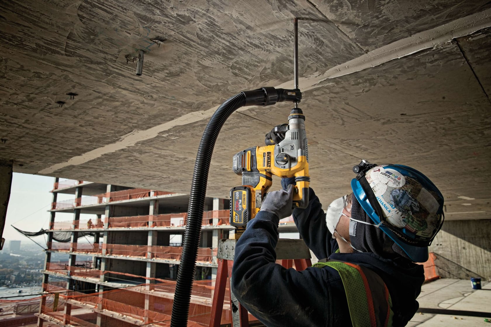 A construction worker wearing a safety helmet and mask uses a DEWALT rotary hammer drill to drill into a concrete ceiling. The worker stands on a ladder and is connected to a dust extraction system. In the background, a multi-story building is under construction.
