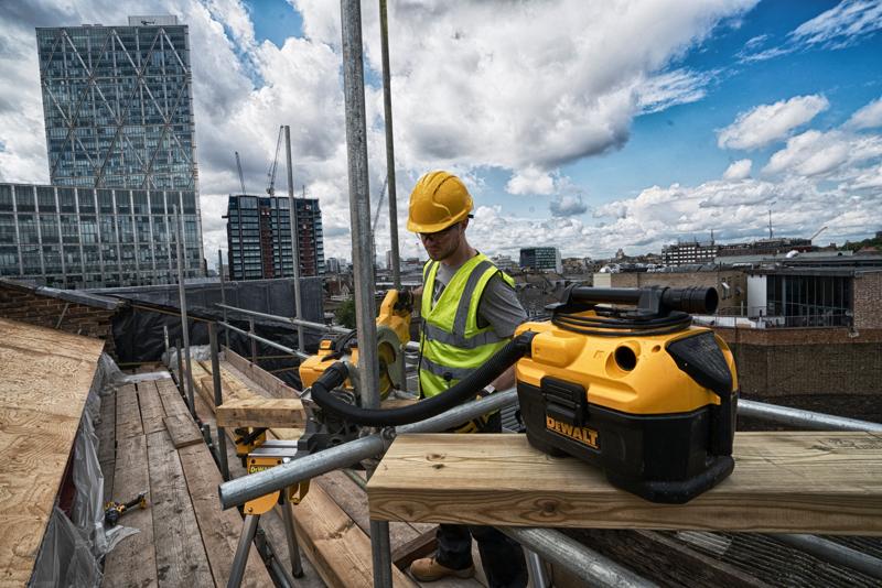 A DEWALT DCV584 portable vacuum cleaner on a construction site, with a worker in a hi-vis vest and helmet operating power tools on scaffolding, and city buildings visible in the background.