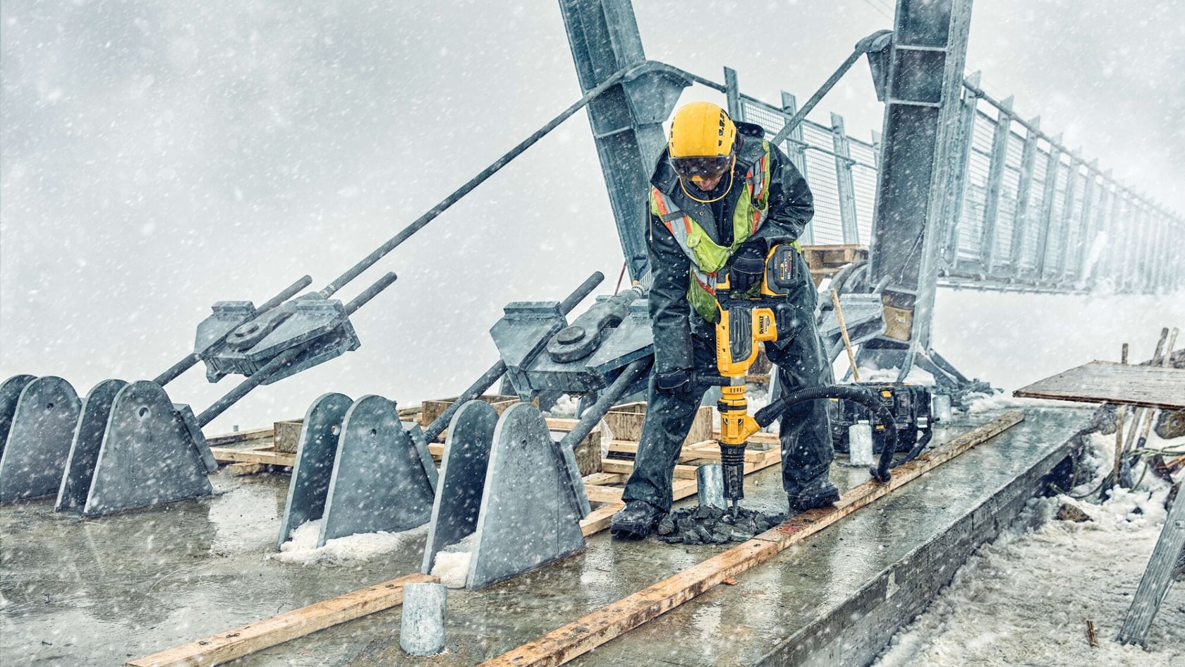 A construction worker wearing protective gear and a yellow helmet uses a DEWALT power tool to drill into concrete on a bridge during snowy weather.