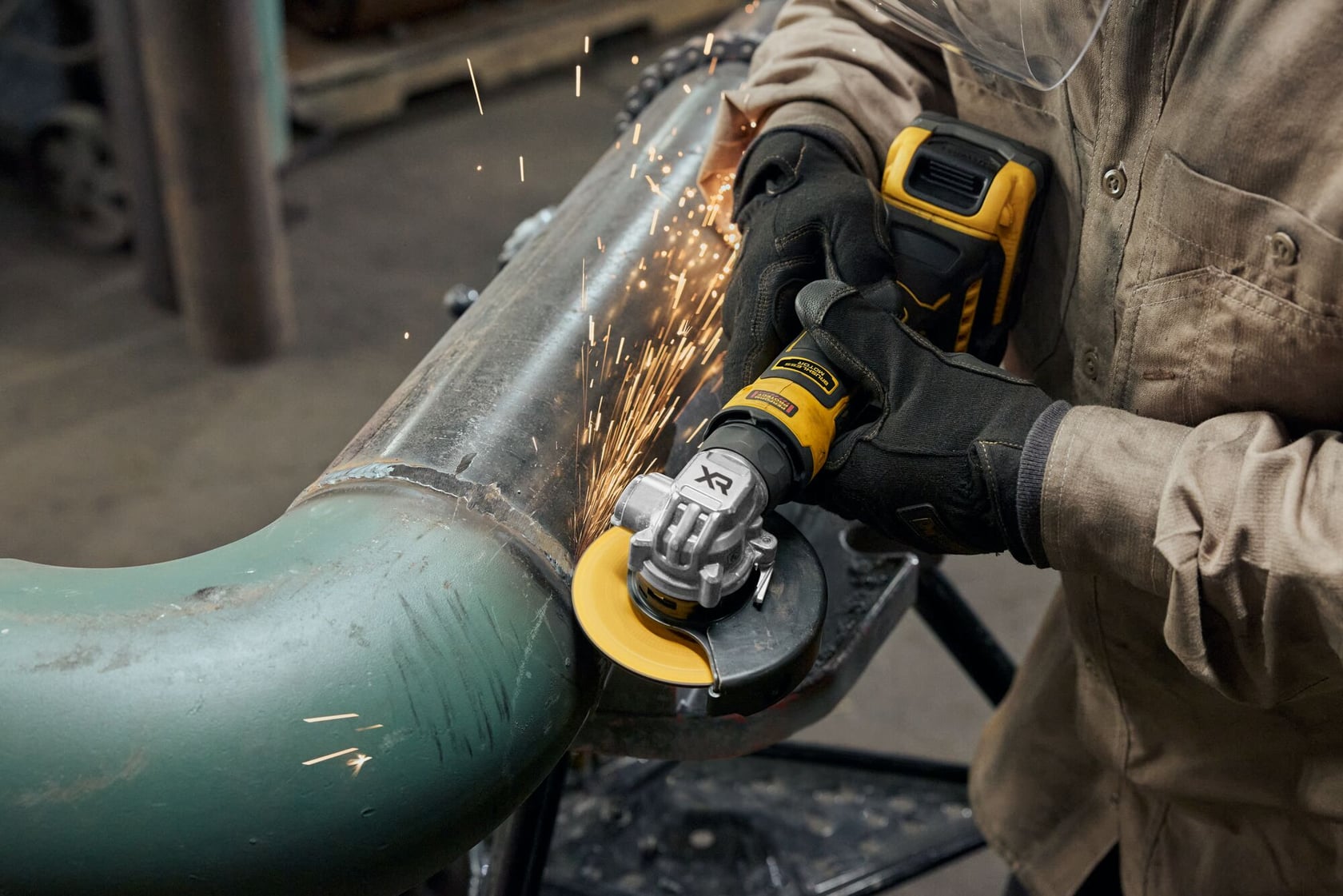 A person wearing protective gloves and a work jacket is using a yellow DEWALT cordless angle grinder to cut or grind a metal pipe, with sparks flying from the tool.