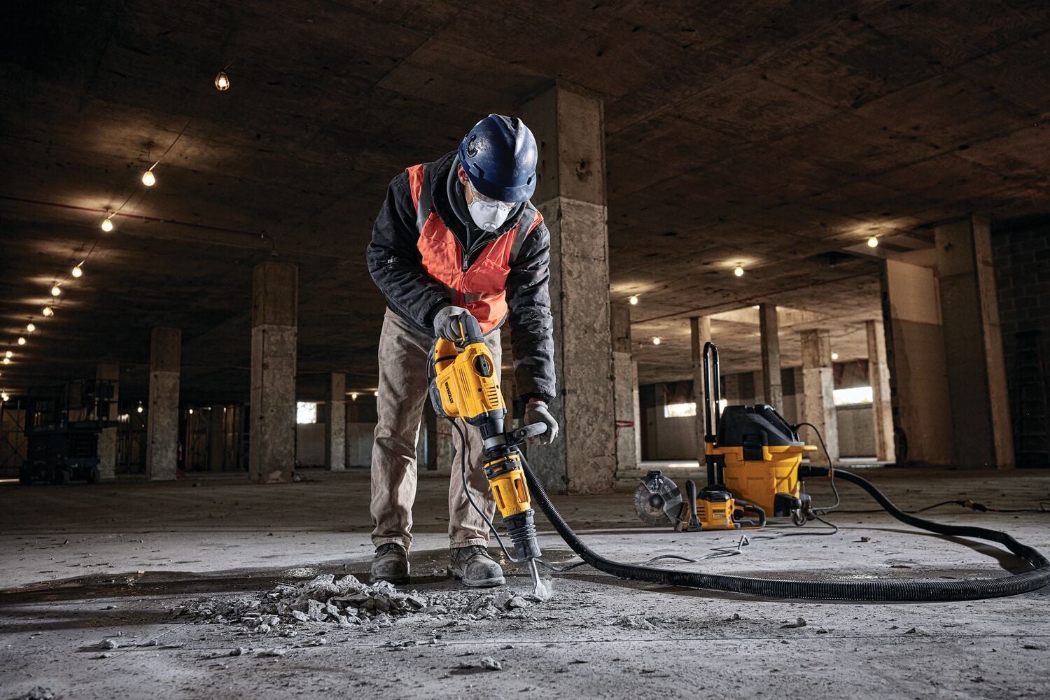 Un trabajador con equipo de seguridad usa un martillo demoledor DEWALT (SKU: D25832K) para romper concreto en el piso de un edificio grande sin terminar. Una aspiradora industrial amarilla está conectada a la herramienta mediante una manguera, visible en el fondo.