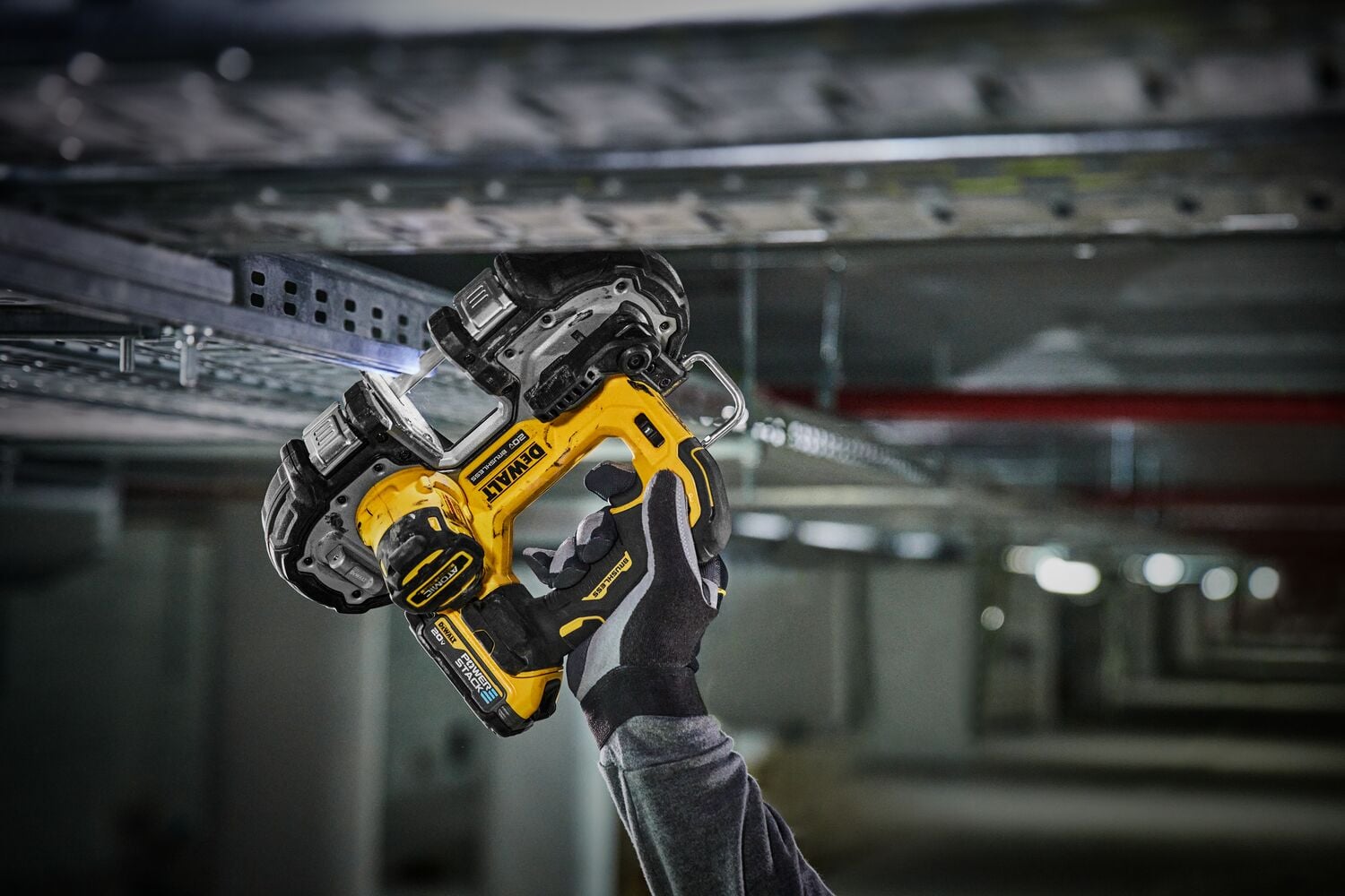 A person wearing gloves is using a yellow DEWALT cordless band saw to cut a metal support beam in an industrial setting.