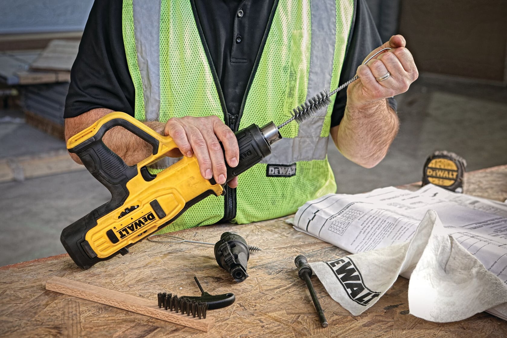 A person wearing a safety vest is using a DEWALT nailer tool with a cleaning brush attached. The tool and brush are on a workbench along with additional DEWALT parts, a tape measure, and papers.