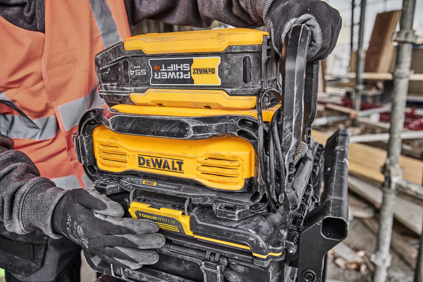 Close-up of a person wearing gloves and a safety vest, handling stacked DEWALT PowerShift 5.5Ah battery and ToughSystem storage units on a construction site.