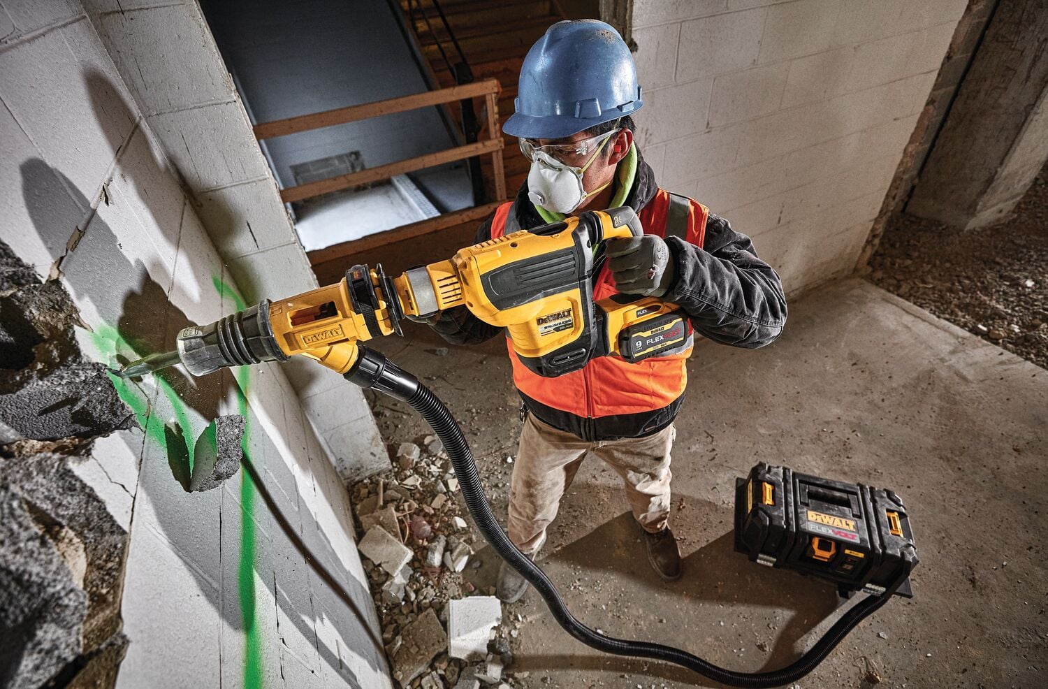 A construction worker wearing a blue hard hat and an orange safety vest uses a DEWALT rotary hammer drill to break through a concrete wall. The tool is connected to a dust extraction system, and a DEWALT tool case is placed nearby on the floor.