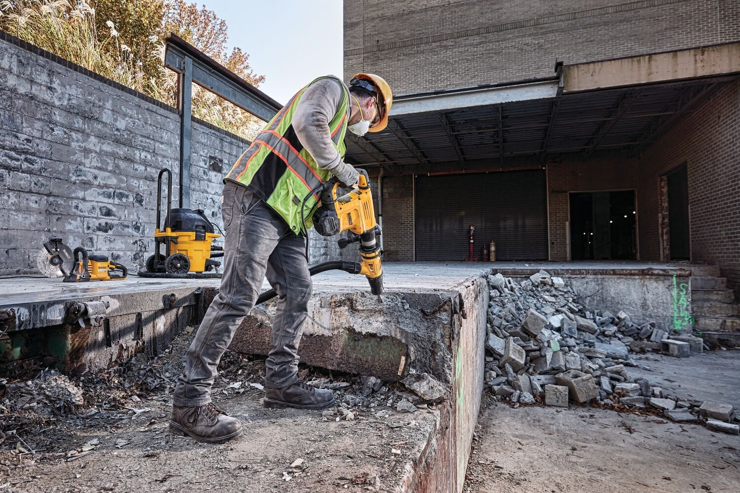 Un trabajador de la construcción que lleva equipo de seguridad y chaleco reflectante utiliza un martillo demolición DEWALT para romper concreto en un sitio de trabajo al aire libre. Se pueden ver otras herramientas y equipos DEWALT en el fondo.