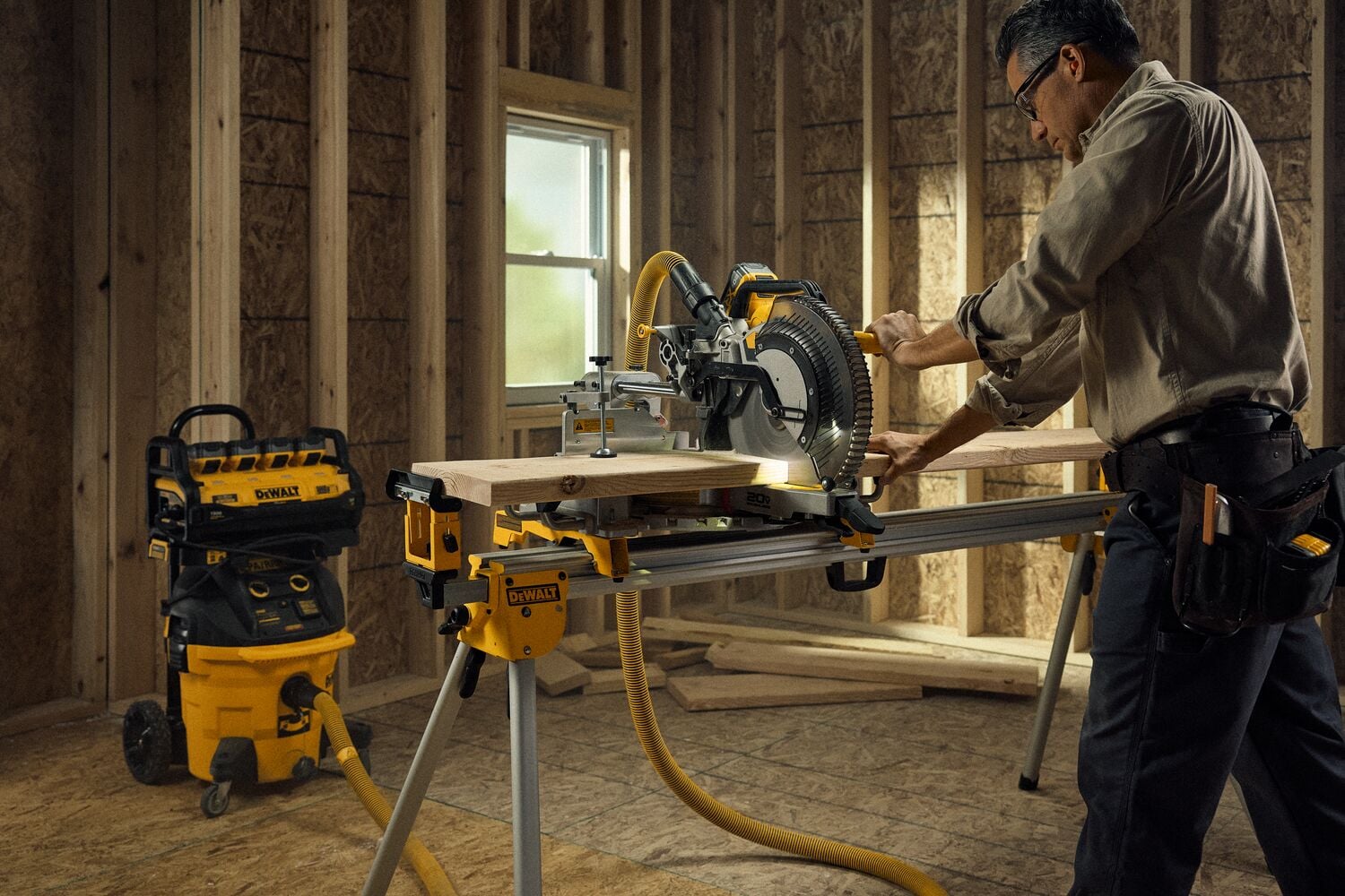 A person using a DEWALT miter saw to cut wood on a stand inside a partially constructed room. There is a DEWALT workshop vacuum and storage system in the background.