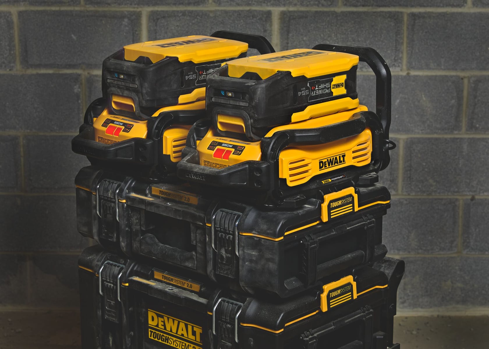 A stack of DEWALT ToughSystem toolboxes with yellow and black DEWALT battery chargers placed on top, set against a grey brick wall.