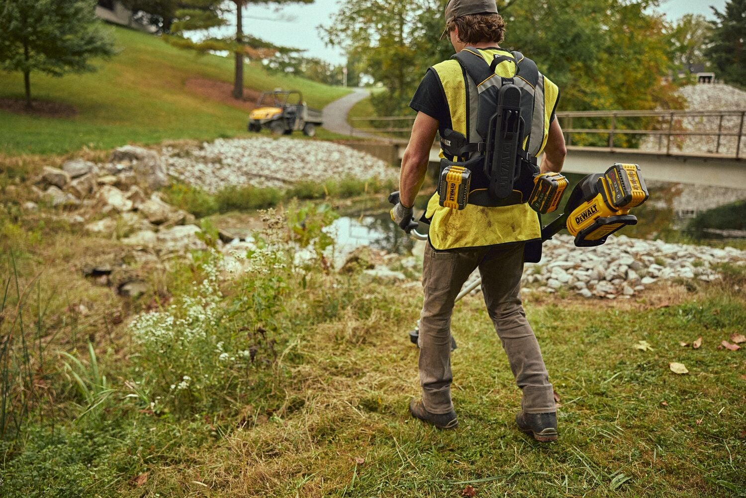 A person wearing a yellow vest and carrying a DEWALT backpack battery system is walking outdoors near a stream, with DEWALT power tool batteries attached to the backpack.