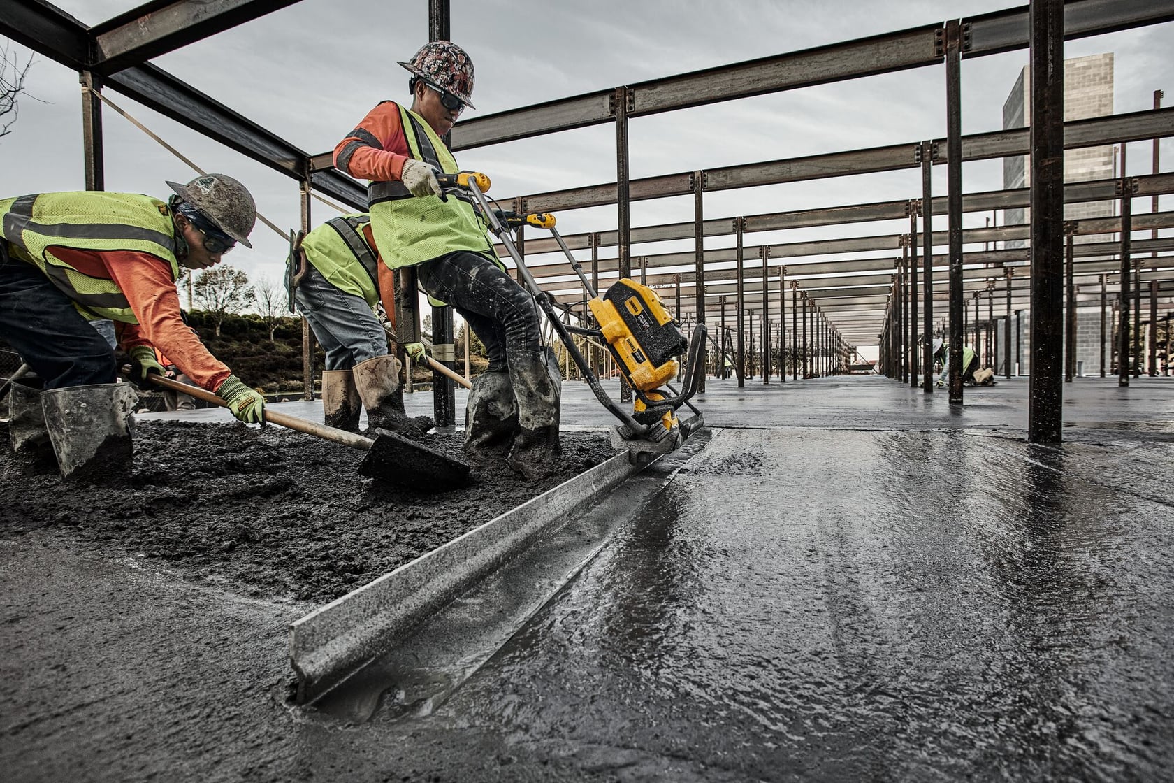 Construction workers in safety gear are using a DEWALT power tool to smooth freshly poured concrete on a large building site with steel beams and framework.