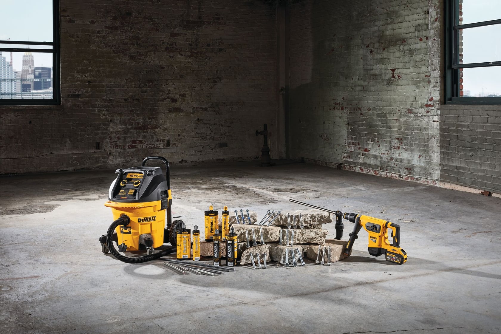 A DEWALT branded set of tools and accessories, including a wet/dry vacuum, an electric power tool, drill bits, and bottles of lubricant arranged on a concrete floor in an industrial space with brick walls and large windows.