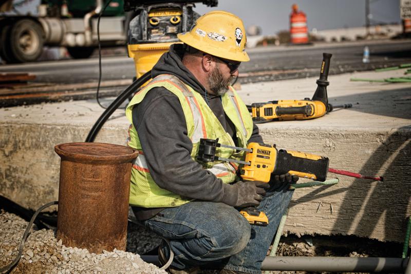 A construction worker wearing a yellow hard hat and safety vest is using a DEWALT DCE595D1 power tool to work on a concrete surface at a construction site. The worker is kneeling next to pipes and equipment, with another DEWALT tool resting on the concrete nearby. The worker's face is blurred for privacy.